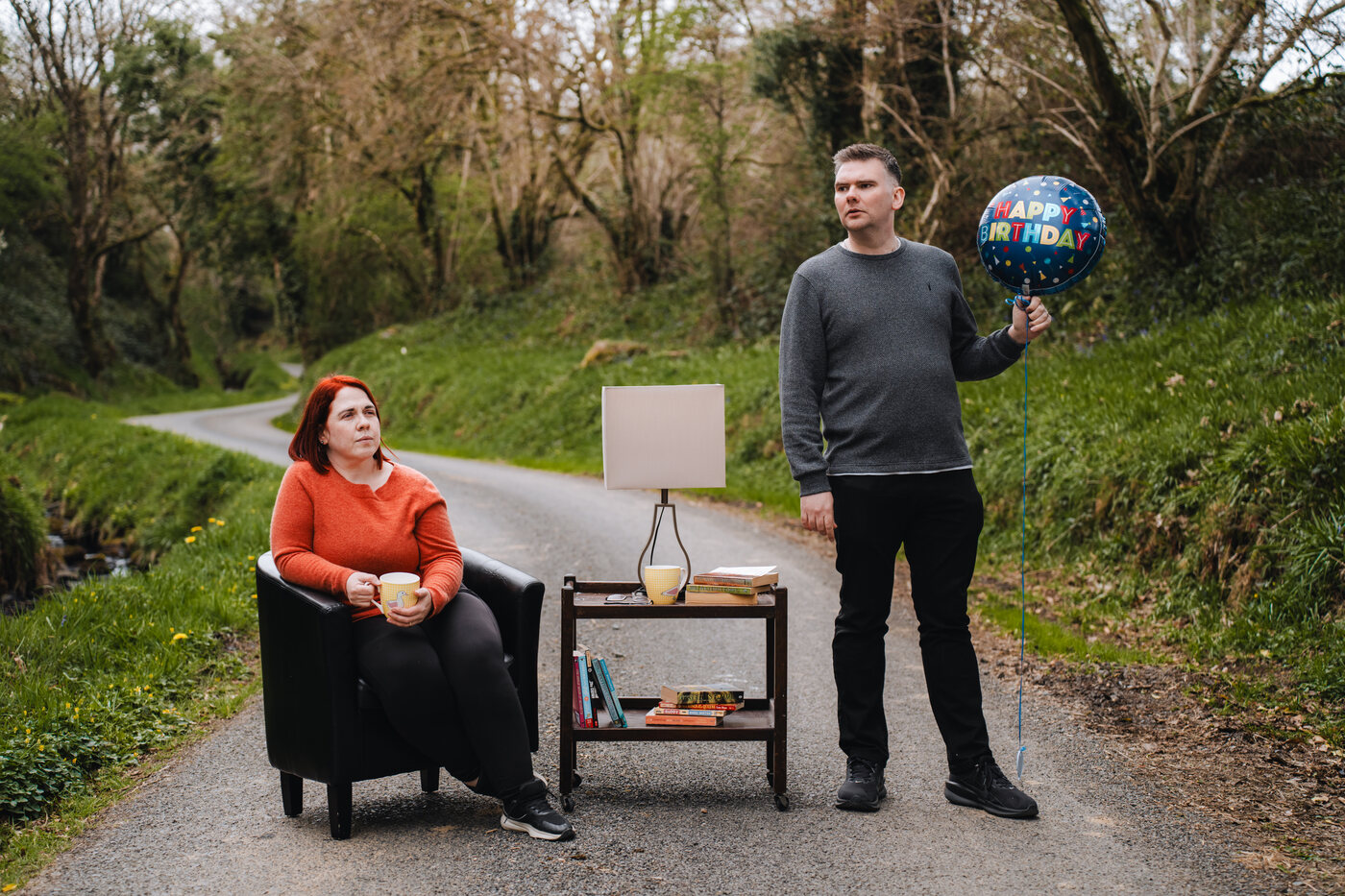 Matthew McMahon and Eileen Gomez in a promotional image for The Flowers of May, with living room furniture arranged on a country road