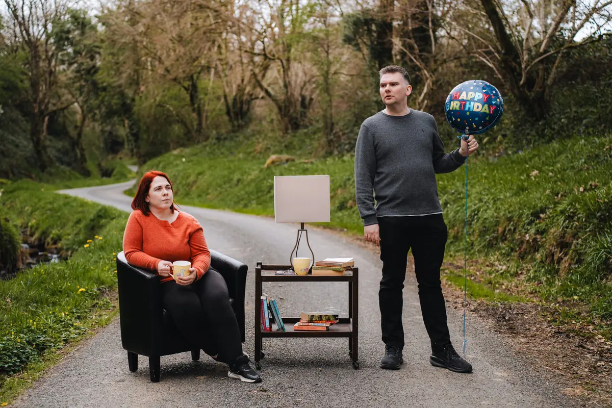 Matthew McMahon and Eileen Gomez in a promotional image for The Flowers of May, with living room furniture arranged on a country road
